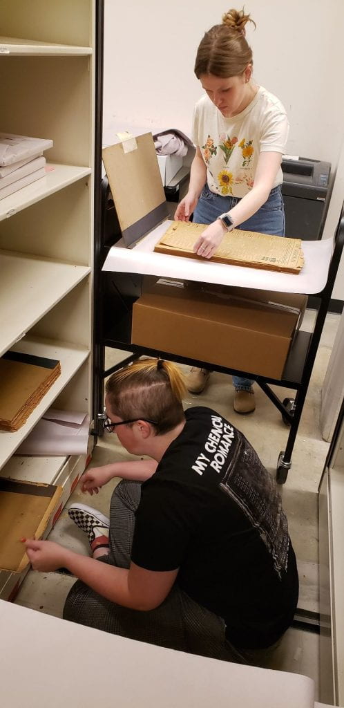One person sits on floor to remove fragile newspapers from compact shelves; one person stands at a book cart to straighten them out.